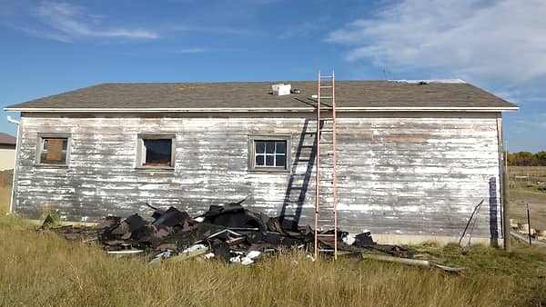 A weathered clapboard building with peeling white paint, an orange ladder to the eave and torn shingles heaped on the grass