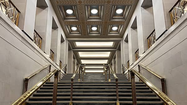 A wide marble staircase seen head-on, with polished brass handrails and a gilded coffered ceiling above