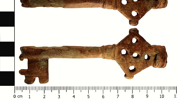 Two corroded iron keys with pierced cloverleaf handles lying on a white ground beside a centimeter scale