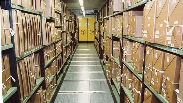 A long storeroom aisle lined with tied cardboard bundles on green metal shelves ending at yellow doors