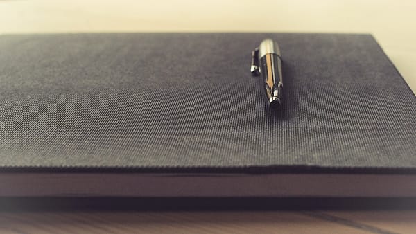 A capped metal pen resting on the closed gray fabric cover of a thick book on a wooden tabletop