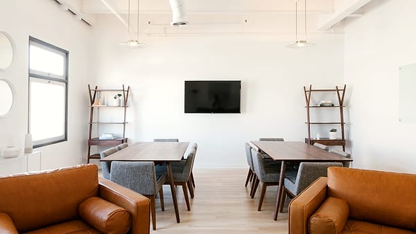 A bright meeting room with two walnut tables, gray upholstered chairs and a wall-mounted flat screen