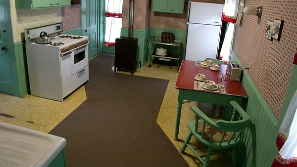 A period kitchen with mint green cabinets, a white gas range, and a red table set with plates and cups