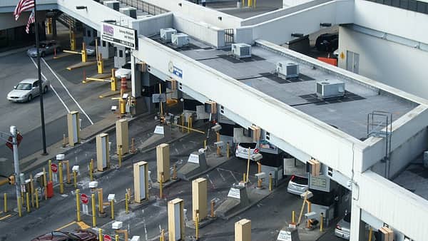 Vehicle lanes at a border inspection plaza, booths under long white canopies with yellow bollards and a flag