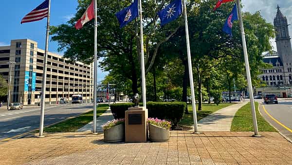 Tall silver flagpoles flying national and state flags over a brick plaza with a small bronze statue on a plinth