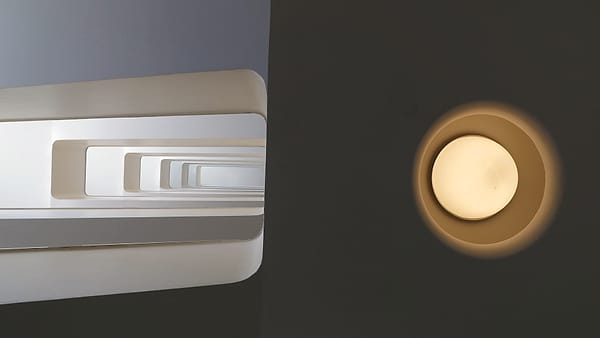 A white rectangular spiral stairwell seen from below, beside a round glowing wall light on a dark wall