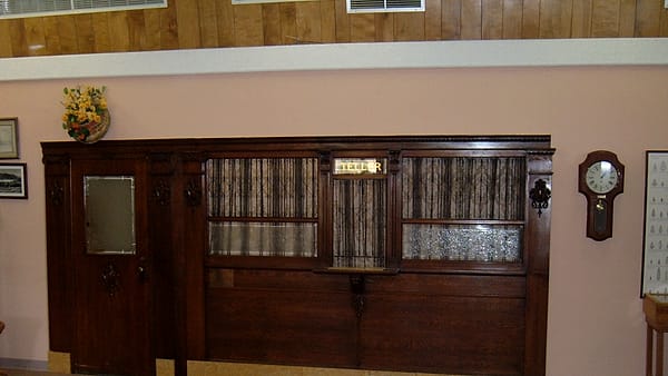 A dark wooden counter with grilled windows and a small lit TELLER sign, set against a pale pink wall