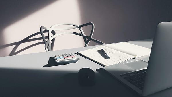 A slim laptop, an open notebook with a pen, and a pocket calculator on a pale desk beside an empty chair