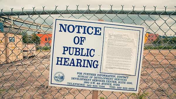 A large white notice board with bold blue lettering fixed behind a chain-link fence at a bare dirt lot