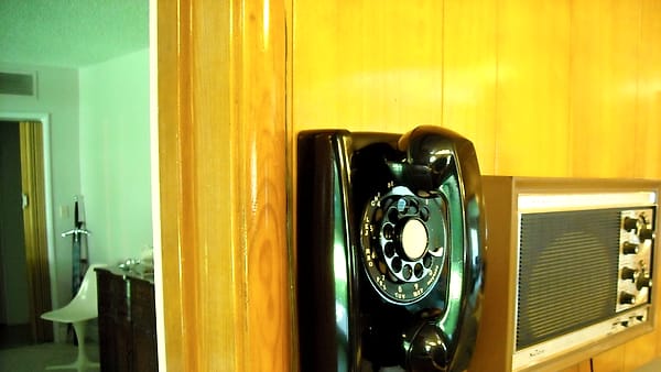 A black rotary dial telephone mounted on knotty pine paneling next to a boxy tabletop radio