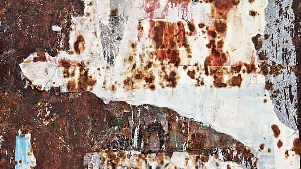 A rusted metal surface streaked with orange corrosion and patched with torn, weathered scraps of paper