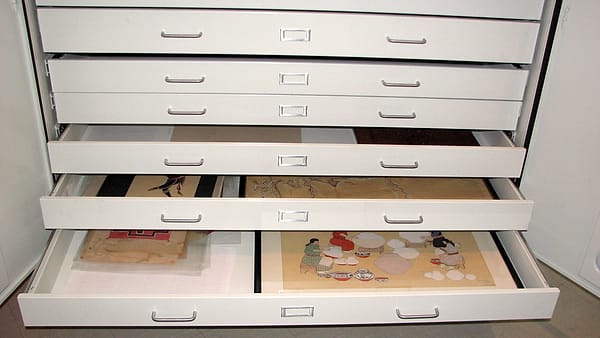 A bank of wide white flat-file drawers pulled open, each holding flat sheets laid out under tissue