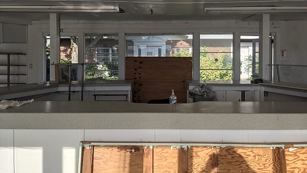 A long laminate counter runs across a stripped room, with bare shelves, plywood sheeting and sunlit windows behind