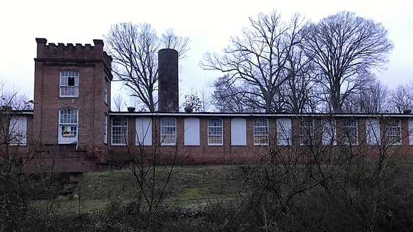 A long brick factory building with rows of tall arched windows, a crenellated tower and a chimney behind bare trees