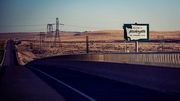A green roadside welcome sign beside a curving highway crossing dry hills strung with power pylons