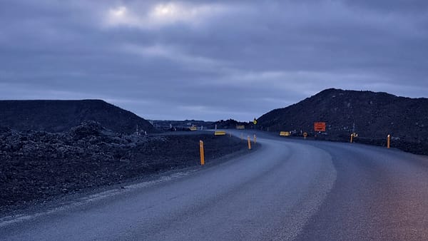 A paved road curving between dark gravel mounds at dusk, marked by orange posts under heavy cloud
