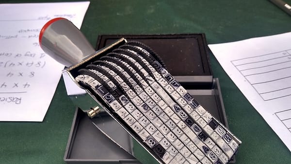 A hand stamp with rows of rotating rubber character bands resting on an open ink pad beside handwritten notes