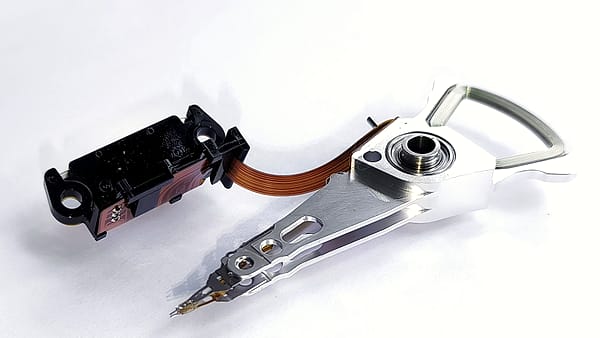 A hard drive actuator arm assembly resting on white, its polished arm and copper ribbon cable exposed