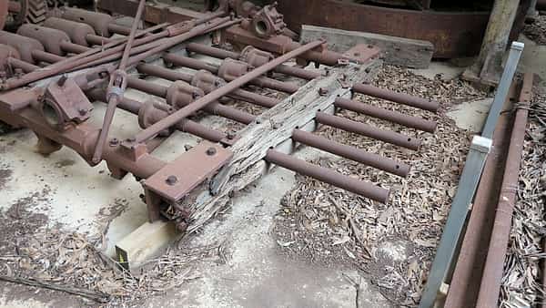 Rusted iron rods and machine frames lying on a concrete floor beside a split timber beam and dry debris