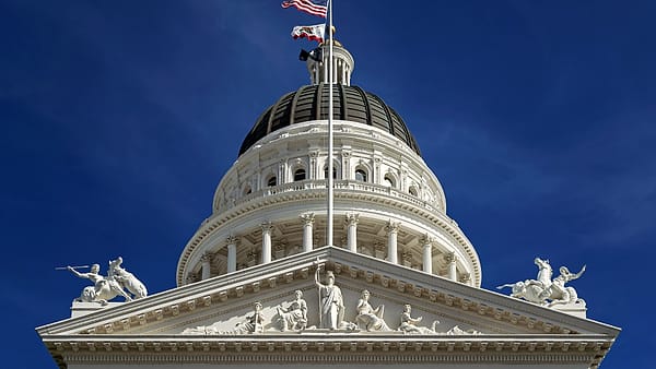 A white domed building with a sculpted pediment and two flags flying above it against a deep blue sky