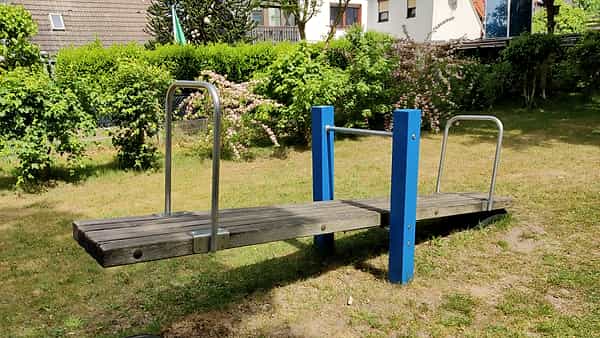 A wooden seesaw plank between two blue posts with metal handles, set on grass in front of trimmed hedges