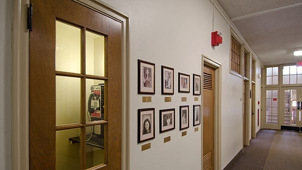 A wooden booth with a wall payphone behind a glazed door, opposite framed portraits along a quiet corridor