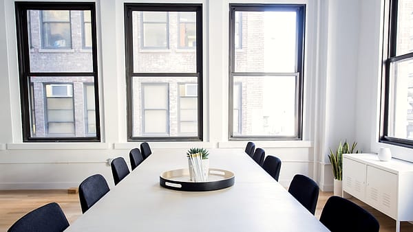 A long white conference table lined with dark chairs beneath three tall black-framed windows overlooking a brick wall