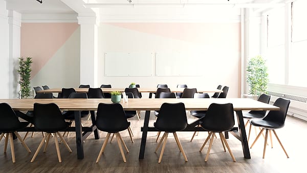 Long pale wood tables ringed by black molded chairs in a bright studio room with whiteboards and potted plants