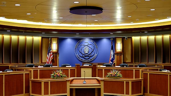 A curved wooden council chamber dais with leather chairs, two flags and flower arrangements below a blue wall