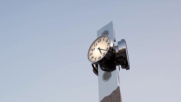 A double-faced public clock mounted on a slim metal post standing against a clear pale blue sky