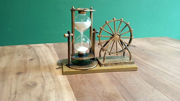 A brass hourglass with white sand beside a small spoked brass wheel on a wooden table against a teal wall
