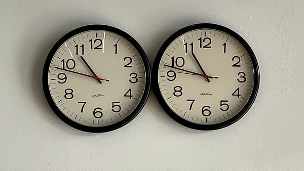 Two identical round black-rimmed wall clocks hung side by side on a pale wall, cream dials with red second hands