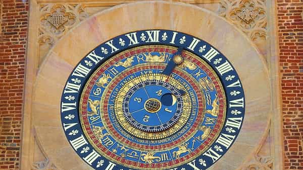An ornate blue and gold astronomical clock dial with Roman numerals and zodiac symbols set in a red brick wall