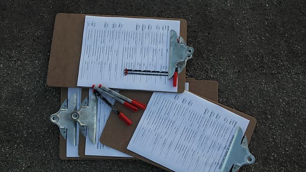 Three clipboards holding printed forms lying on gray asphalt with red-capped marker pens resting across them