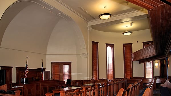 A high-ceilinged paneled hall with wooden benches, a raised desk and two flags standing beside an arched wall
