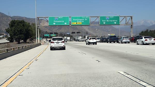 Traffic on a wide freeway passing beneath large green overhead direction signs, with hazy mountains ahead