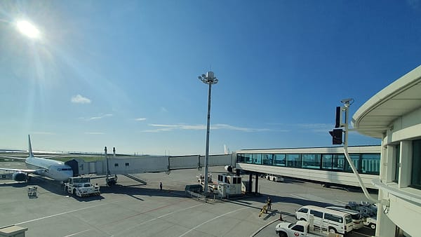 A white airliner parked at a jet bridge on a sunlit apron, with service trucks and a tall floodlight mast