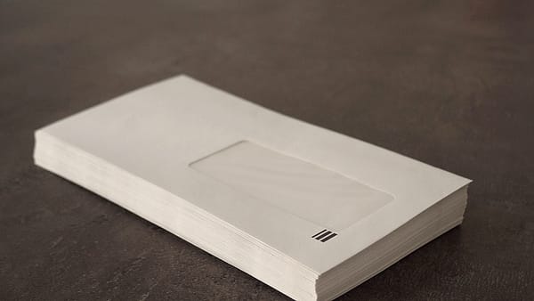 A thick stack of plain cream window envelopes resting on a dark textured tabletop, lit softly from the left