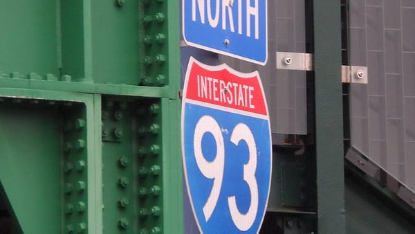 A blue and red interstate route shield below a north marker, bolted to a green riveted steel girder