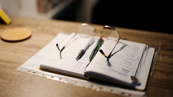 An open ruled notebook on a wooden desk, two pens and wire-rimmed glasses on its pages, a round coaster nearby