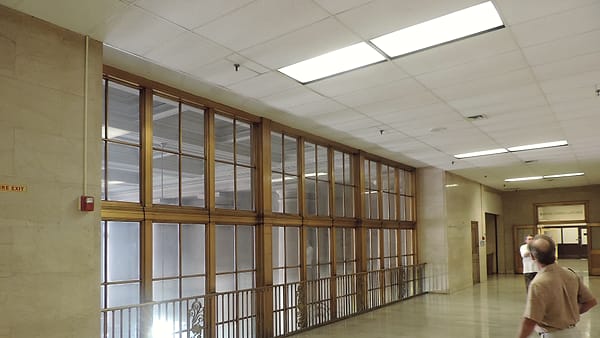 A wide polished corridor lined with tall wood-framed interior windows under a paneled ceiling of fluorescent lights