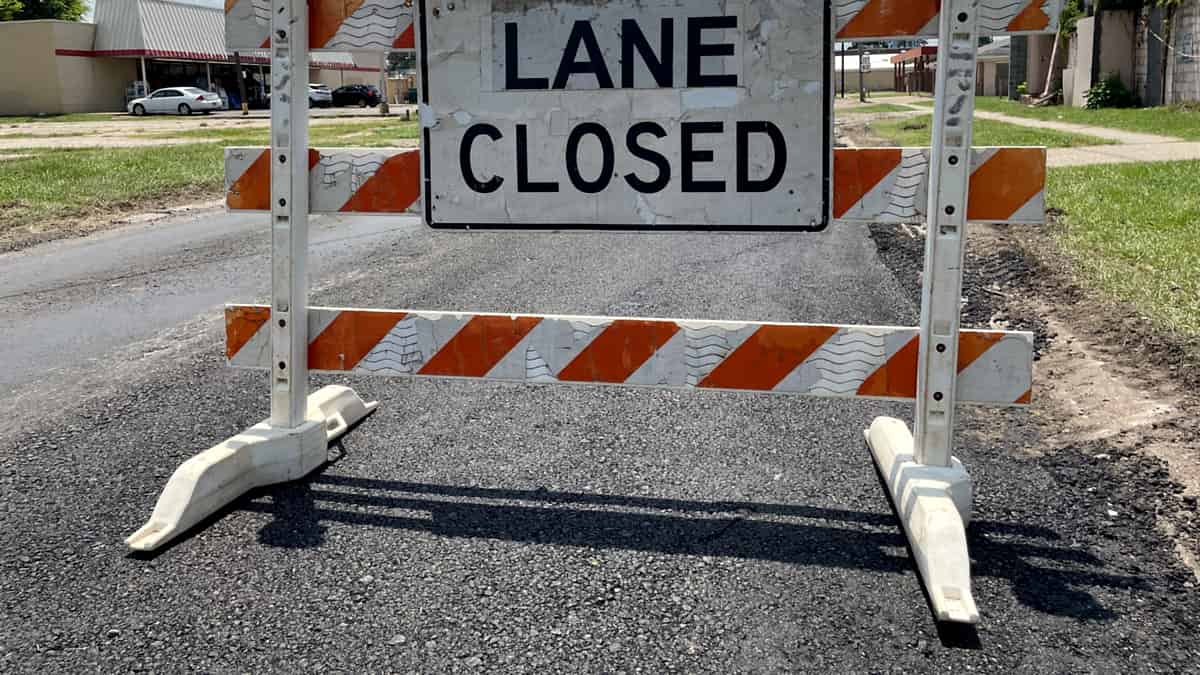 An orange and white striped barricade standing across a paved road under a sign reading LANE CLOSED