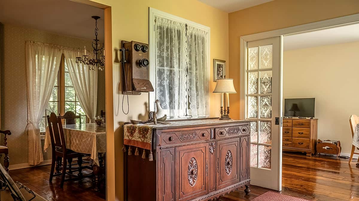 A black wall-mounted crank telephone on yellow plaster beside a lace-curtained window and carved sideboard