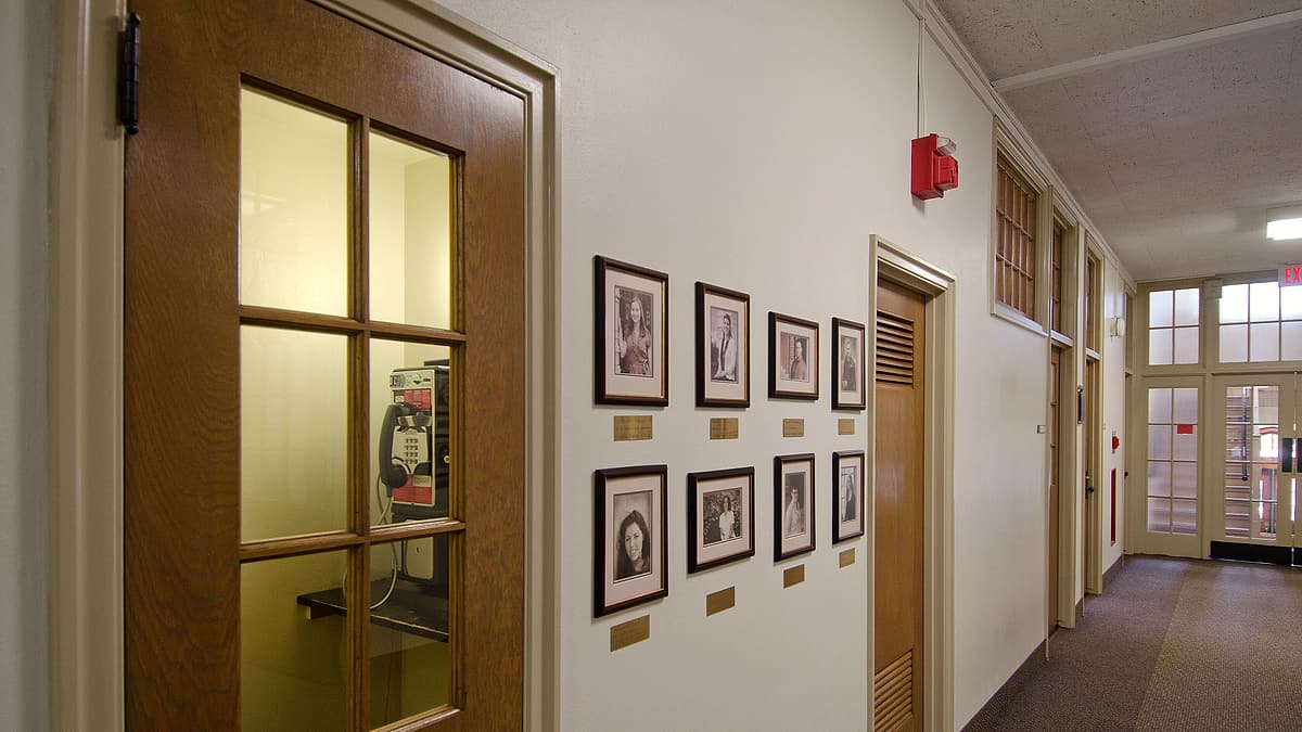 A wooden booth with a wall payphone behind a glazed door, opposite framed portraits along a quiet corridor