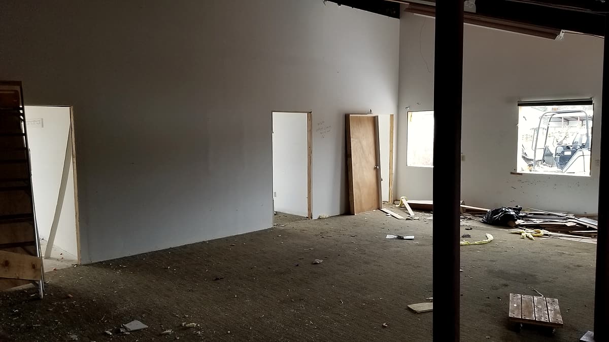 A gutted interior with bare drywall, an unhung wooden door leaning by a doorway and debris on the floor