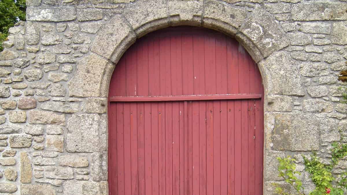 A red painted plank door under a stone arch, set in a rough gray granite wall with ivy at one side